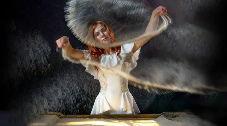 Sand artist for weddings in a white dress creates detailed drawings over an illuminated light table during a live performance.
